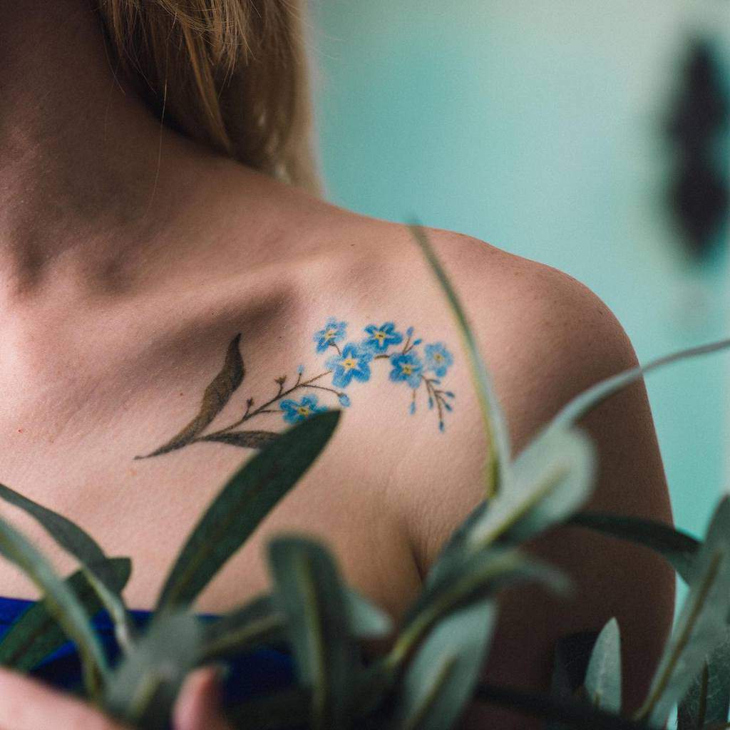 A shoulder tattoo of forget-me-not flowers in a soothing blue, set elegantly above lush green leaves