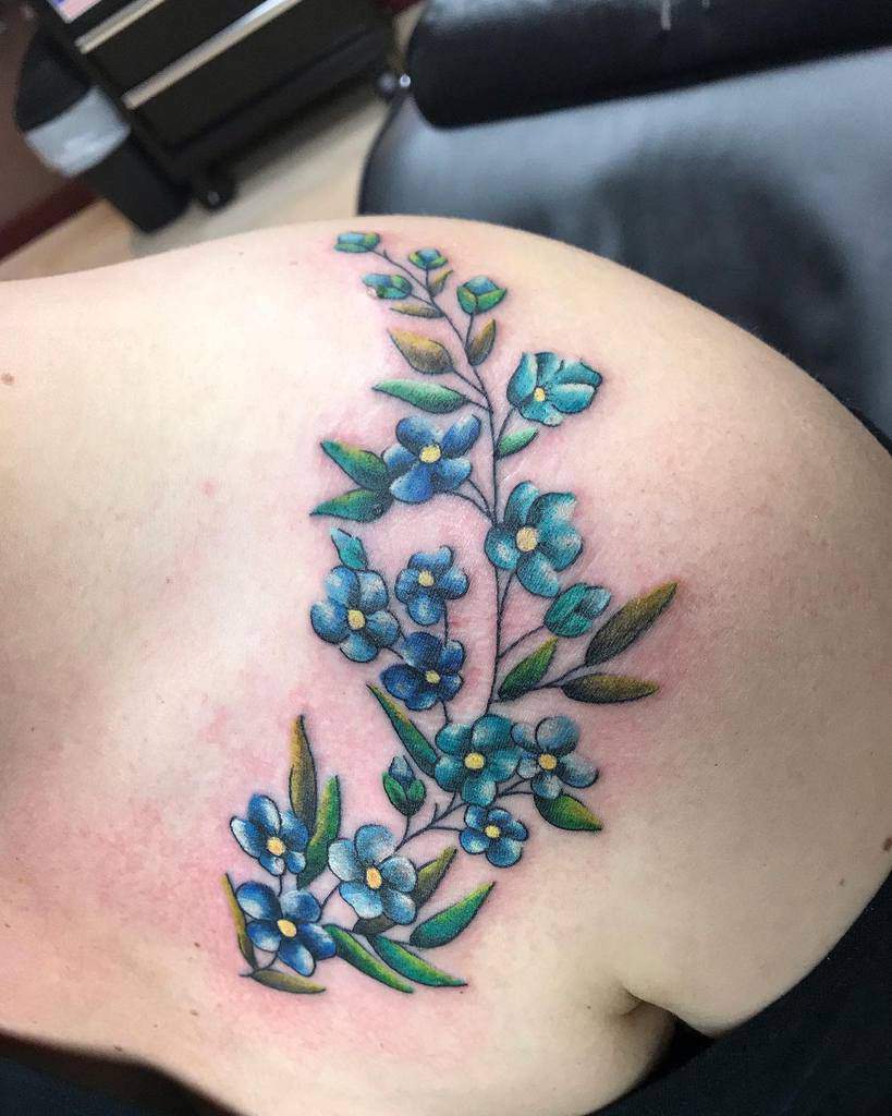 A floral tattoo featuring delicate forget-me-nots with blue flowers and lush green leaves graces the person's shoulder