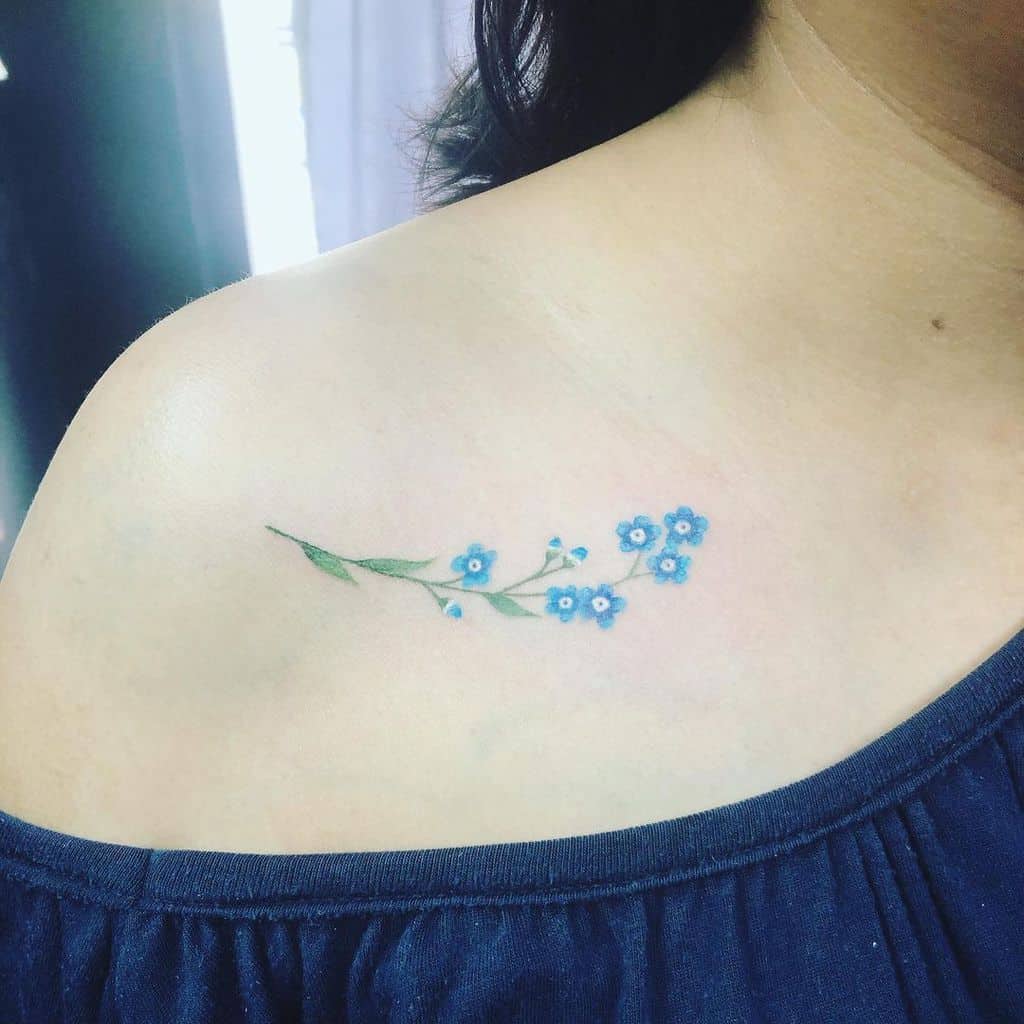A delicate forget-me-not tattoo graces a woman's upper shoulder, showcasing its serene blue petals