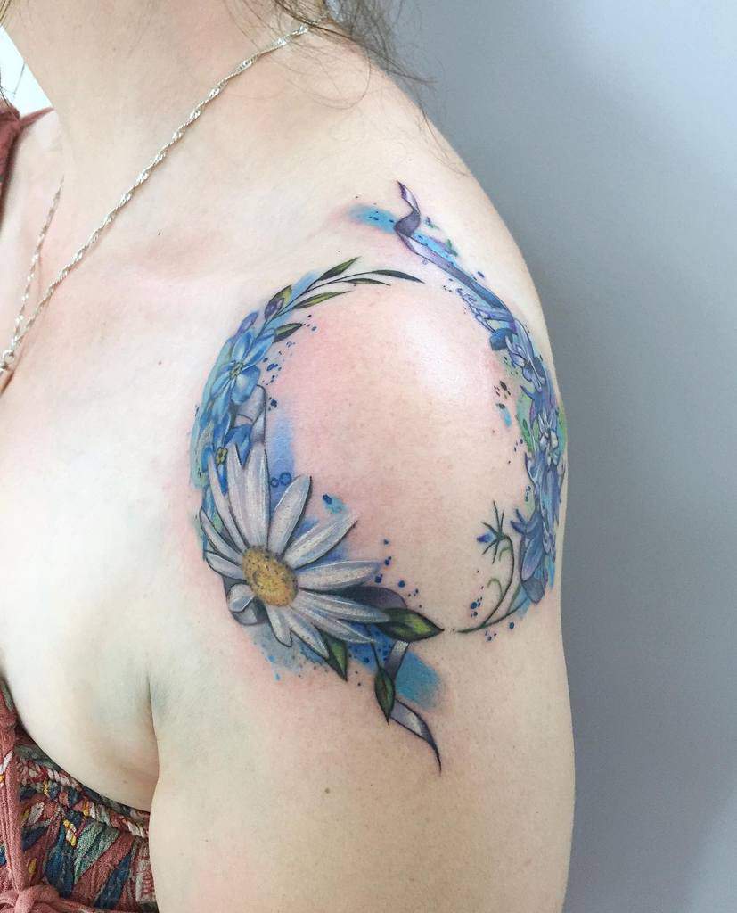 A colorful, watercolor-inspired tattoo of forget-me-not flowers, elegantly wrapping around the shoulder with a daisy accent
