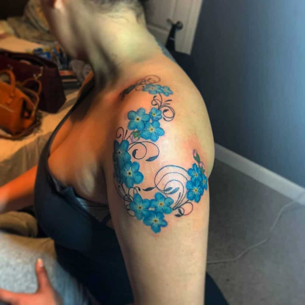 A vibrant tattoo of forget-me-not flowers wrapping around the shoulder, showcasing their detailed blue petals and swirling vines