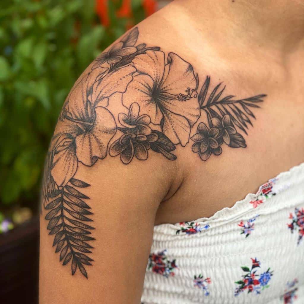 Detailed black and grey shoulder tattoo featuring hibiscus and plumeria flowers with fern leaves, extending across the shoulder and collarbone area