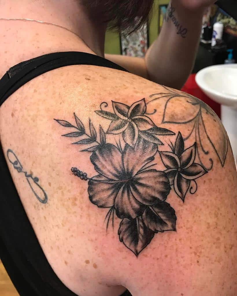 Black and grey shoulder tattoo featuring a hibiscus flower and two plumeria flowers, surrounded by leaves with shading details