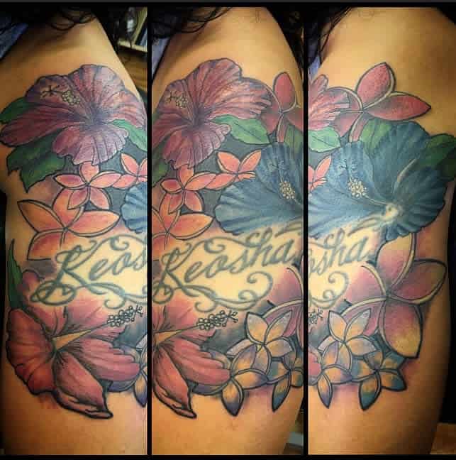 Colorful upper arm tattoo featuring a mix of large hibiscus and plumeria flowers in shades of pink, blue, and orange, surrounding the script 'Keosha' in an elegant font