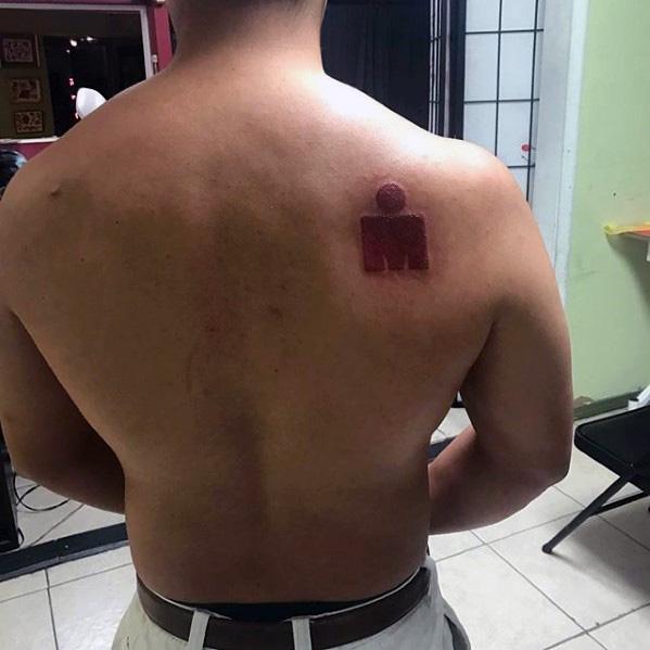 Simple solid red Ironman logo tattoo placed on the upper back near the right shoulder blade, minimalistic with no added elements.