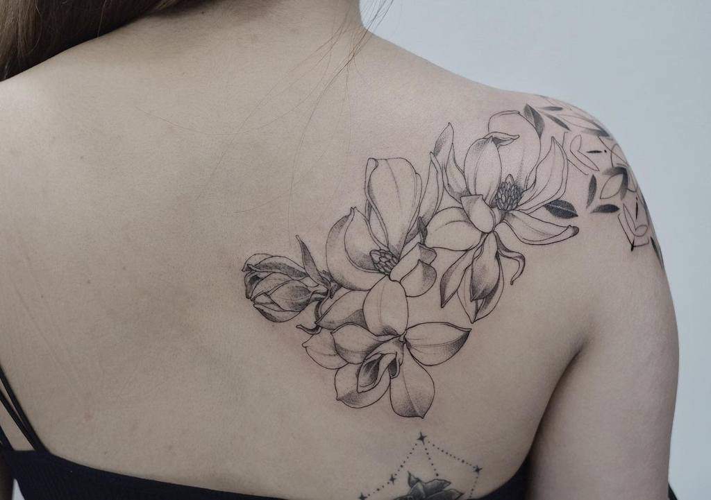 A graceful magnolia tattoo adorns a woman's shoulder and upper back in elegant grayscale, capturing nature's delicate beauty