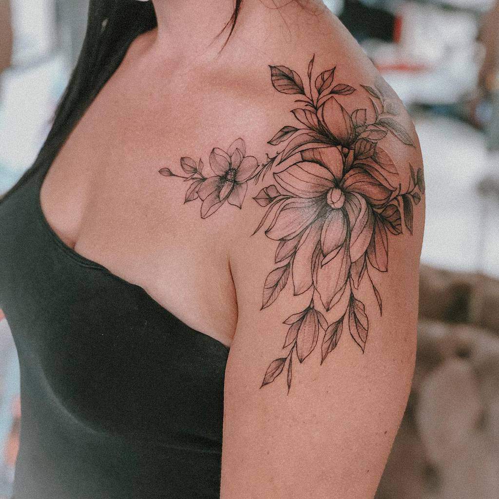 A delicate magnolia tattoo blossoms on a woman's shoulder, surrounded by intricate black leaves and petals