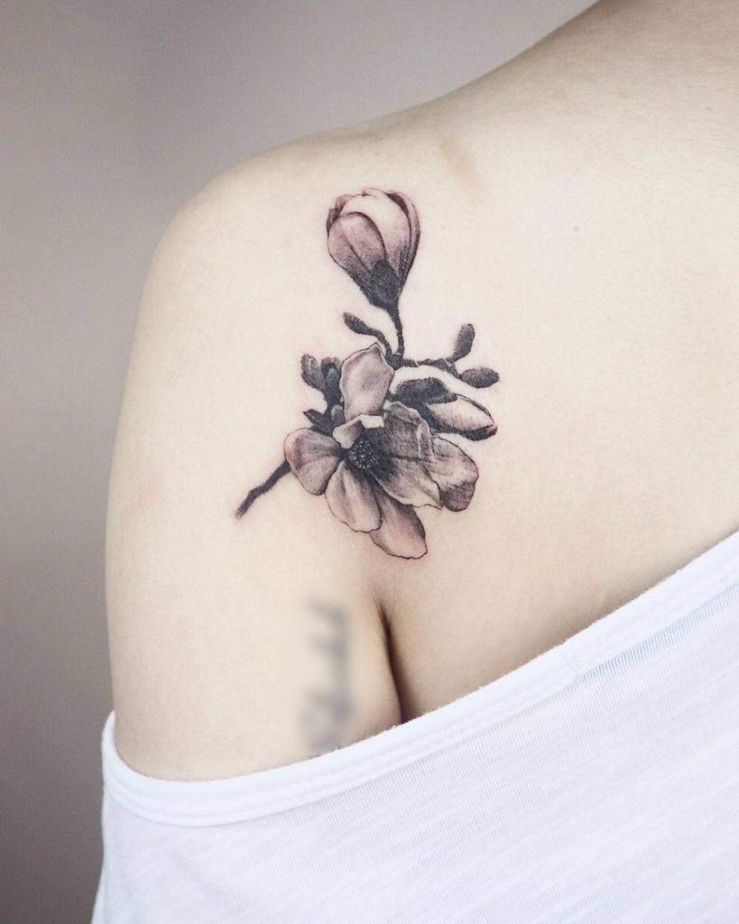 A black and gray magnolia tattoo graces a person's shoulder, its delicate petals beautifully rendered in ink