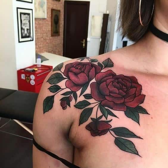 A tattoo featuring a vibrant red rose surrounded by luscious green leaves adorns a woman's shoulder and upper chest