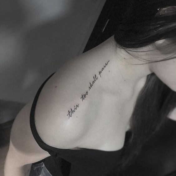 A tattoo on the shoulder that says "this too shall pass" in a cursive font.