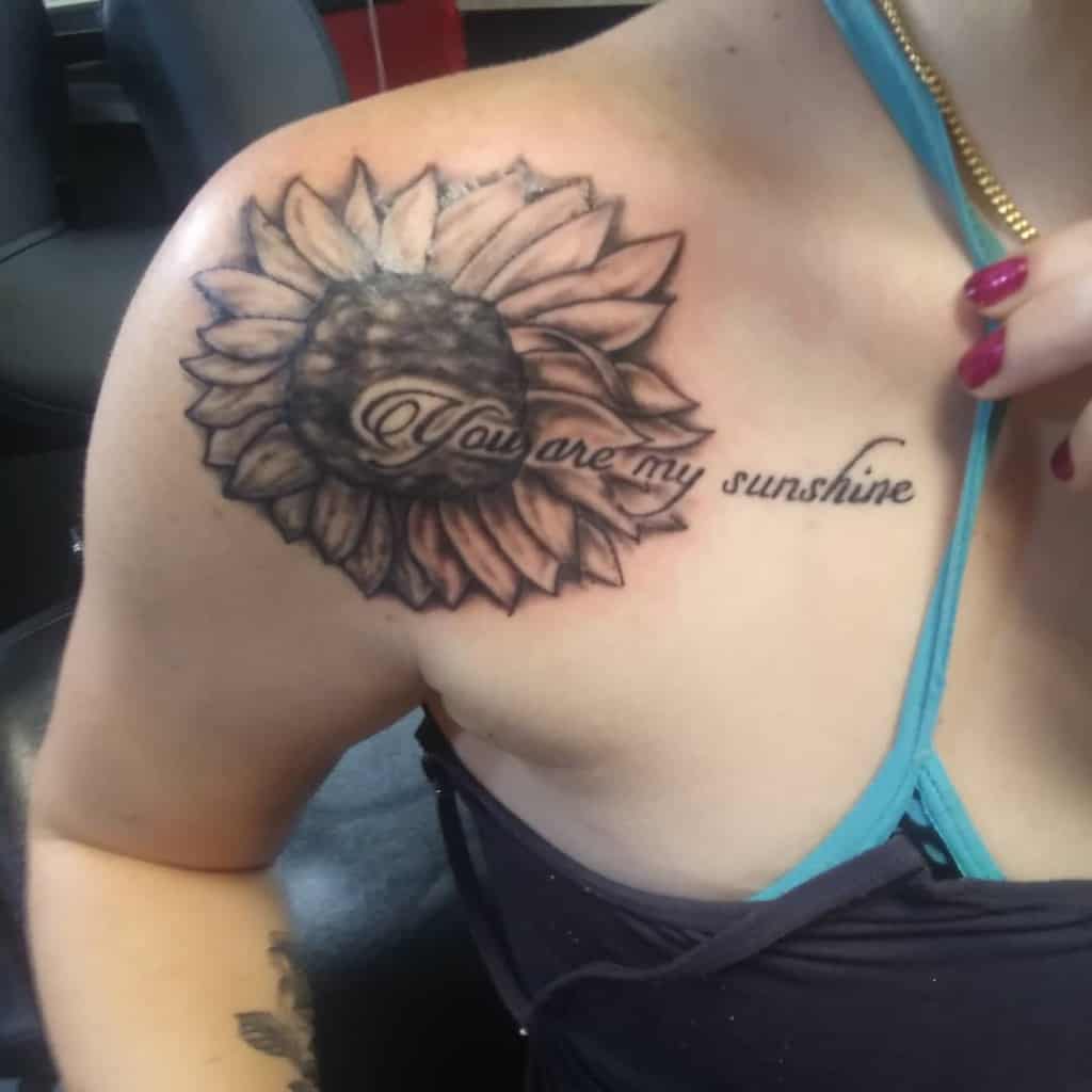 A sunflower tattoo on the shoulder featuring the text "You are my sunshine" without a dot at the end