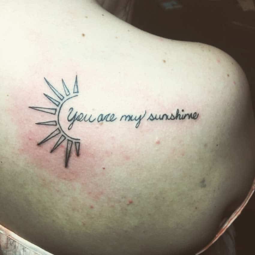 Simple shoulder tattoo with 'You are my sunshine' in script and a minimalist half-sun design in black ink