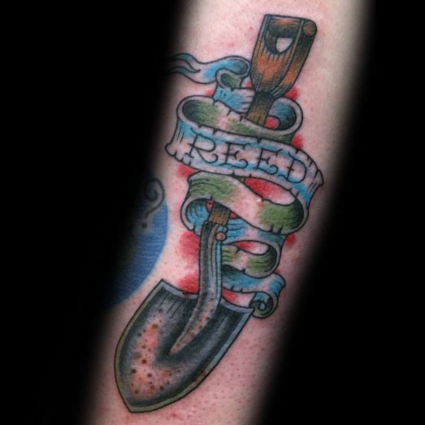 Shovel tattoo with a weathered blade wrapped in a flowing ribbon that reads "GREED," symbolizing struggle, ambition, or consequences of excess.