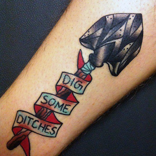Tattoo of a rugged shovel wrapped in a red ribbon with the phrase "Dig Some Ditches," symbolizing hard work, perseverance, and determination.