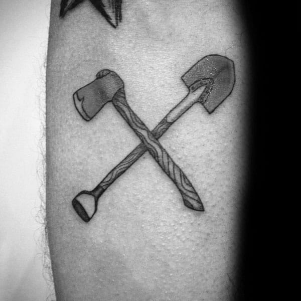 Black and grey tattoo of a crossed shovel and axe with wood grain detailing, symbolizing strength, labor, and resilience in a bold design.
