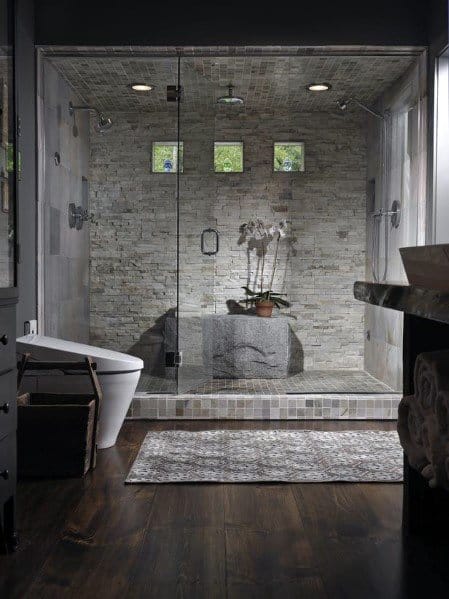 Spacious master bathroom shower with natural stone walls, multiple showerheads, and soft overhead lighting, complemented by unique window accents