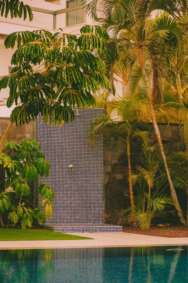 Poolside outdoor shower with blue mosaic tiles surrounded by lush tropical trees and plants.