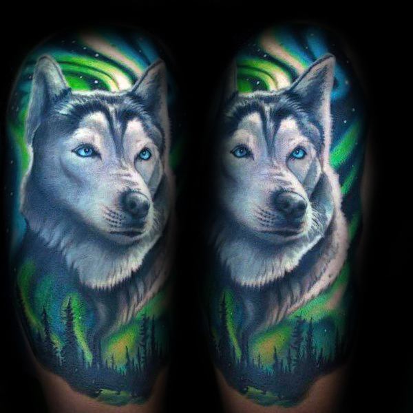 Realistic Siberian Husky tattoo on a man's arm, featuring piercing blue eyes, detailed fur, and a vibrant Northern Lights background for a mystical look.
