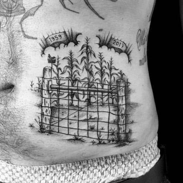 Black and gray tattoo of a cornfield behind a rustic wire fence, featuring memorial dates '1959 - 2017' in a detailed and sentimental design.