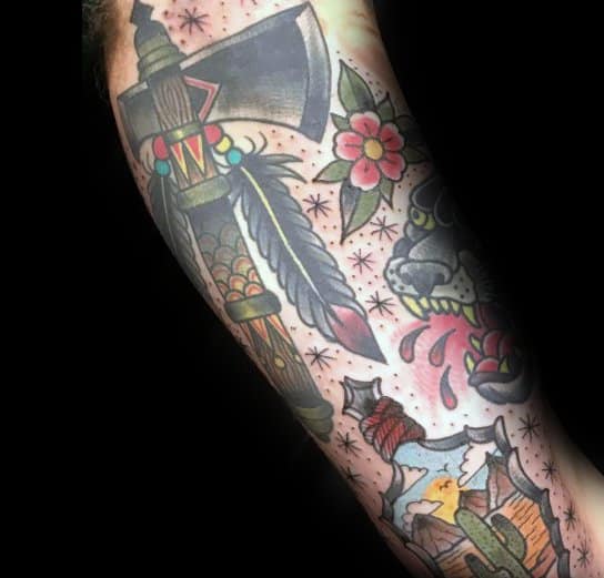 Colorful traditional-style hatchet tattoo on the forearm, featuring intricate feather and bead details, floral elements, and bold shading for a striking look.