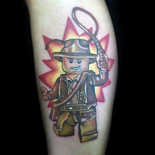 Bold and colorful tattoo of LEGO Indiana Jones, featuring his iconic fedora, whip, and satchel, set against an explosive background.