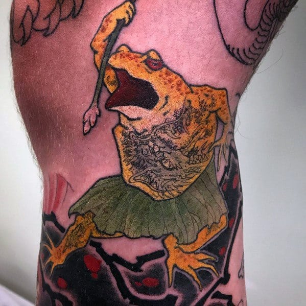 Japanese-style tattoo featuring a fierce orange frog in a warrior pose, wearing a green robe, gripping a staff, and set against a bold traditional background.