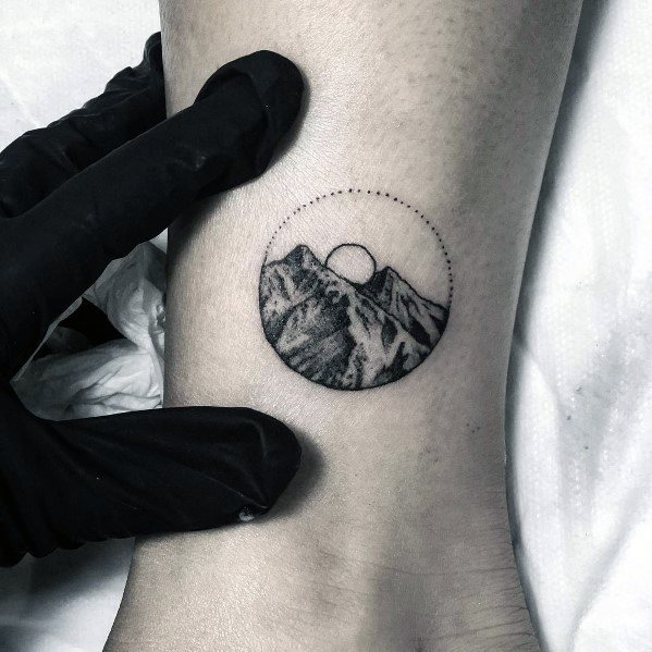 Minimalist circular mountain tattoo on the ankle, featuring fine shading, a rising sun, and a dotted outer frame for a detailed yet subtle design.