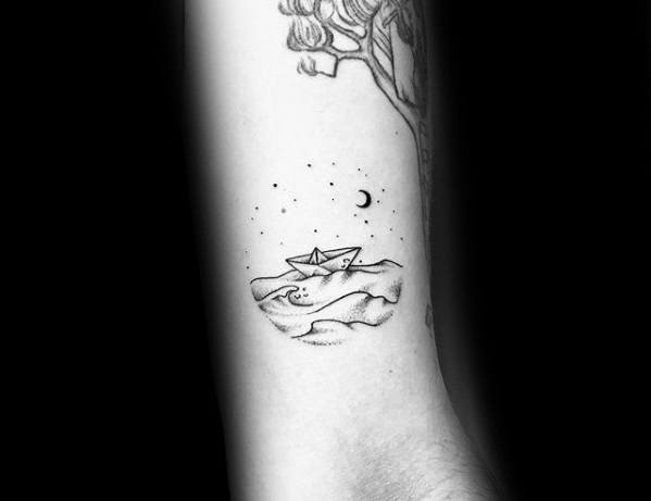 Delicate black ink tattoo of a paper boat on waves under a starry night sky with a crescent moon, symbolizing dreams and life's journey.