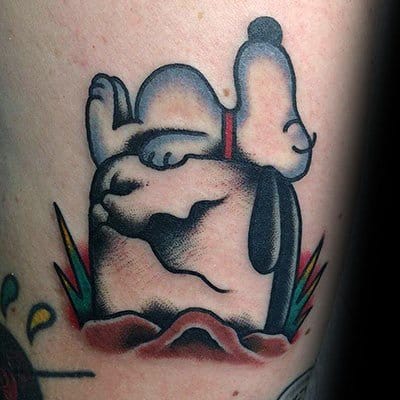 Colorful tattoo of Snoopy peacefully sleeping on a shaded rock, surrounded by grass, with a red collar, placed on a person's arm.