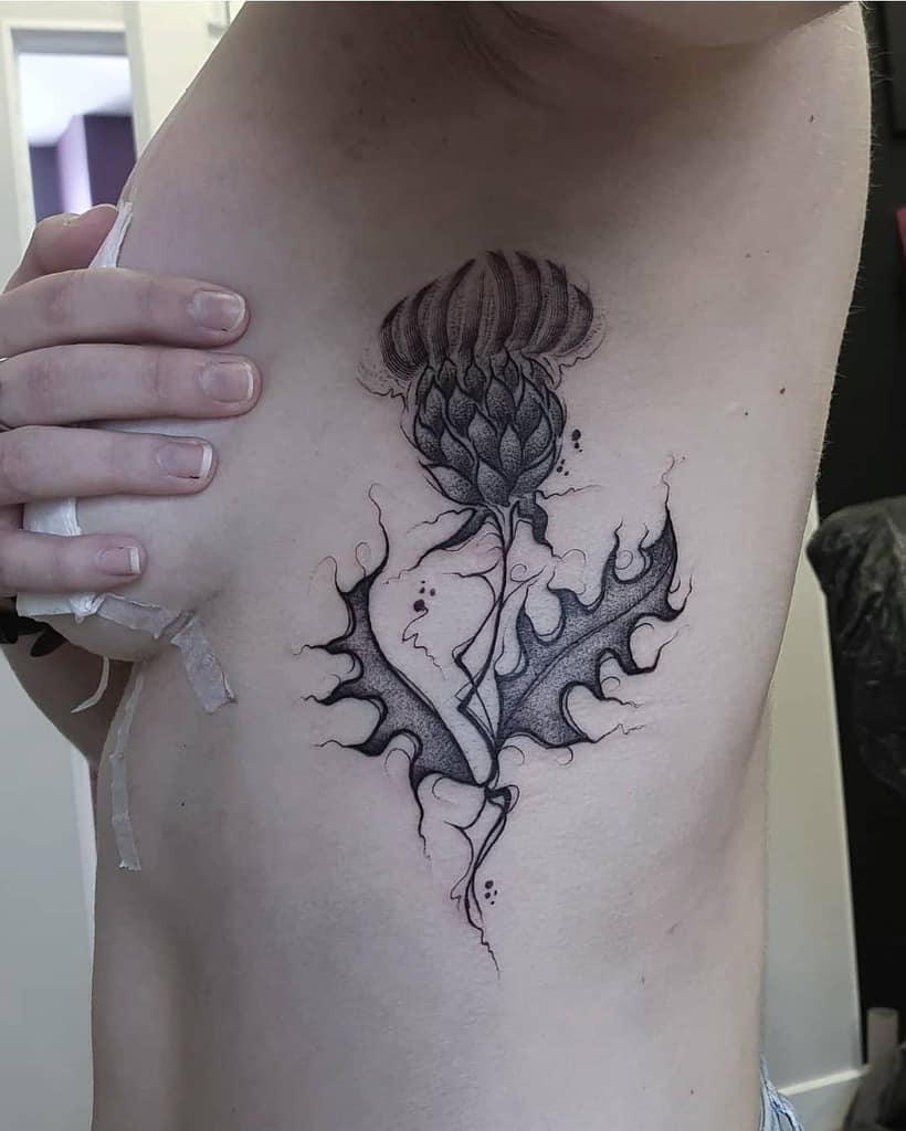 A delicate black and gray tattoo gracefully depicts a thistle, beautifully etched on a woman's side, capturing its intricate details