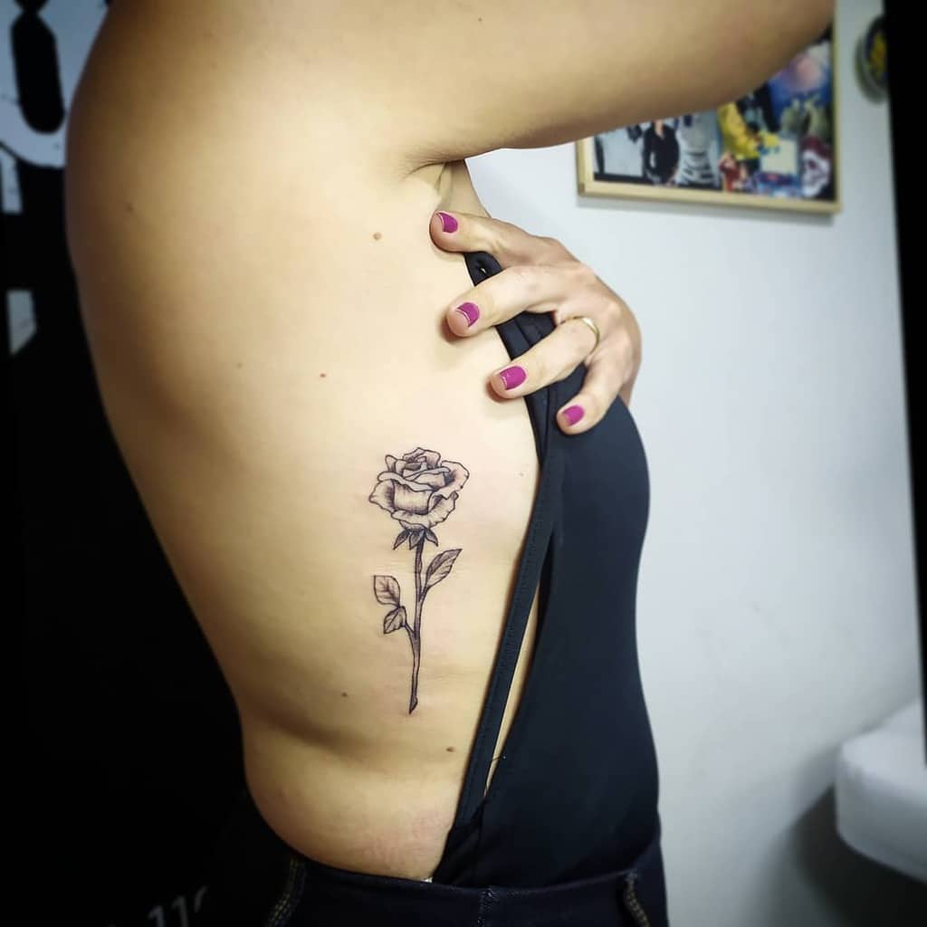 A delicate black and white tattoo of a rose on the side of the torso, featuring fine lines and a minimalistic design