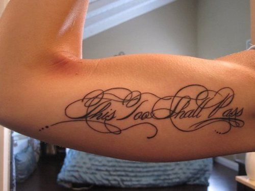 An elegant "This too shall pass" tattoo in ornate cursive script, placed on the inner bicep, representing strength and perseverance.