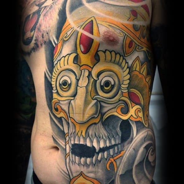 Golden Tibetan skull tattoo on the side torso with wide white eyes, red gemstone crown, and swirling grey smoke accents.
