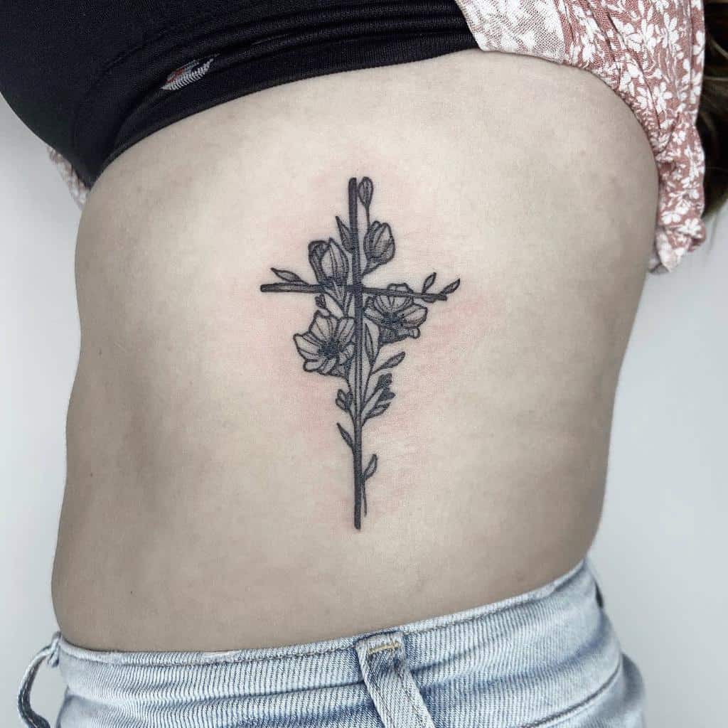 A floral tattoo featuring a detailed black ink cross intertwined with flowers and leaves graces a woman's ribcage