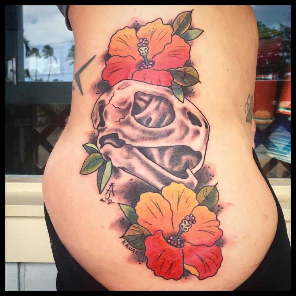 Side tattoo featuring a turtle skull in the center, framed by two colorful hibiscus flowers in orange and red, with green leaves.