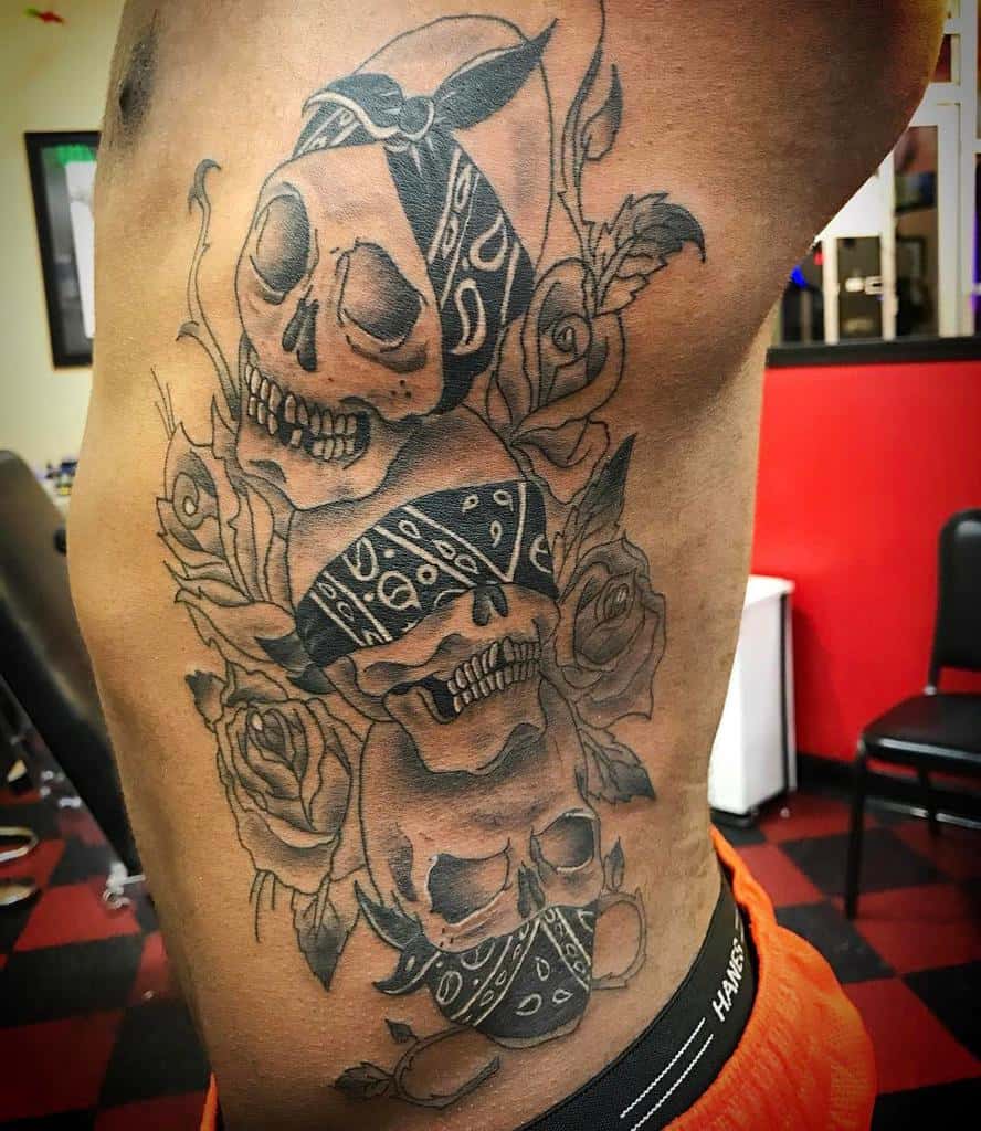 Side tattoo of three skulls wearing bandanas, representing 'Hear No Evil, See No Evil, Speak No Evil,' surrounded by roses and detailed shading