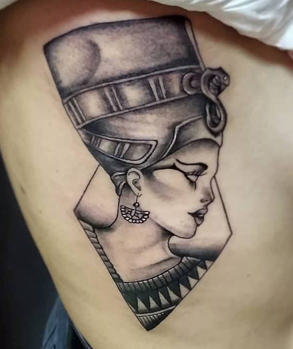 Tattoo of an Egyptian queen with a patterned headdress and an earring, in black and gray ink, on skin