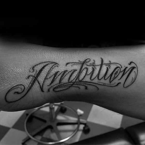 Stylized "Ambition" tattoo graces an arm, set against a backdrop of a chair and tiled floor