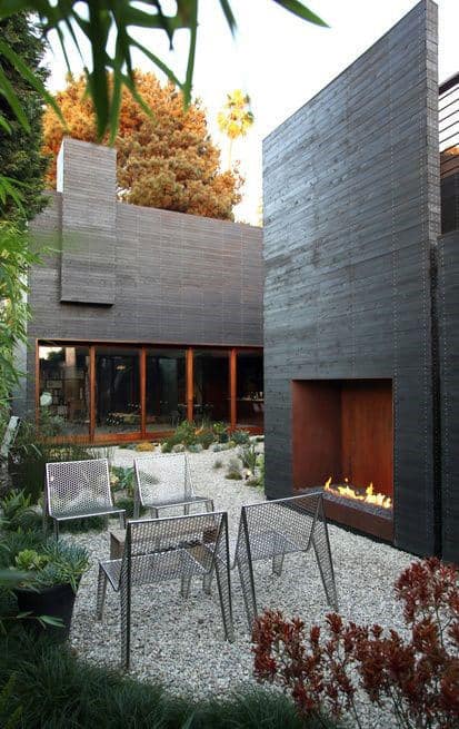 Modern patio with metal chairs around a fireplace, surrounded by greenery and a dark wood-paneled building