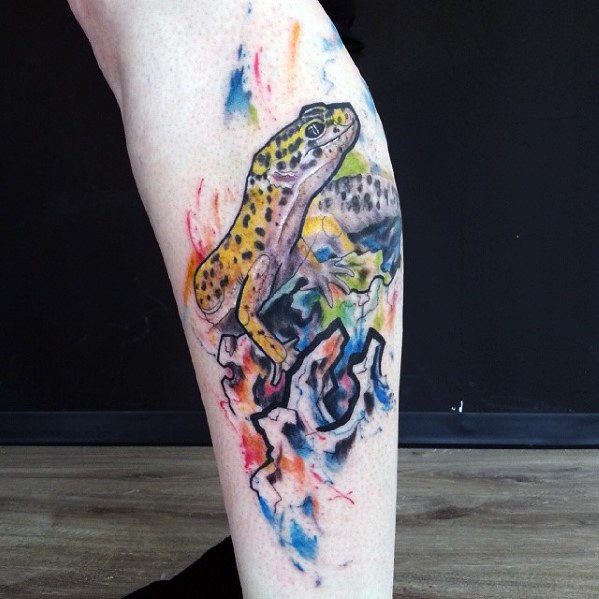 Leopard gecko tattoo on leg with vivid yellow spots and watercolor-style background in blue, red, and orange tones over rocky shapes.