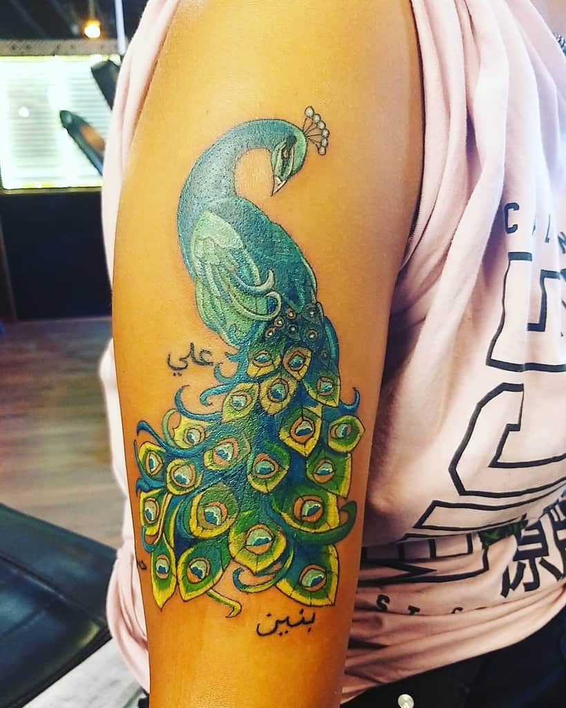 Vibrantly colored peacock tattoo on the upper arm with green and blue feathers, along with Arabic text.