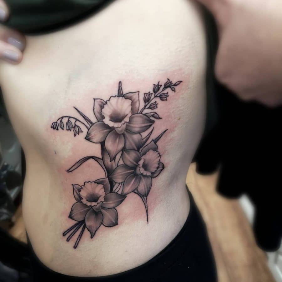 This tattoo is a beautifully shaded floral design featuring several daffodils. The arrangement gives a natural flow across the side of the body, with the flowers appearing vibrant and lively despite the black and grey ink. The shading and placement create a harmonious look.