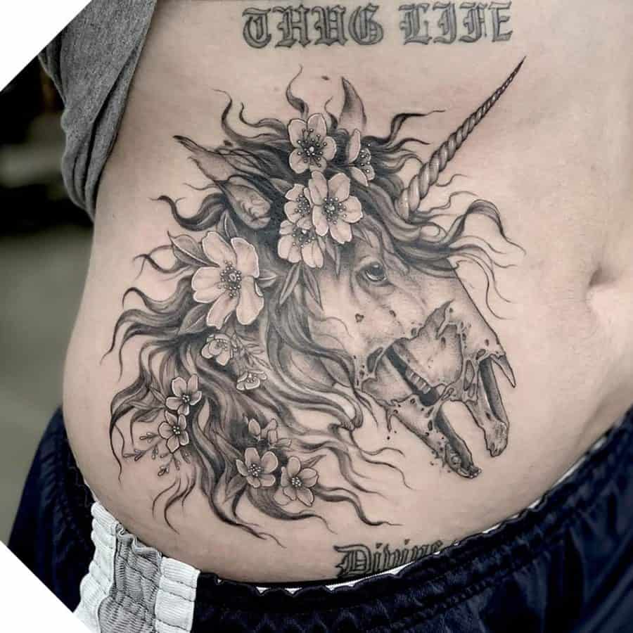 Detailed black and grey tattoo of a unicorn with a decayed, skeletal face, adorned with flowers and flowing hair, blending beauty with darkness.