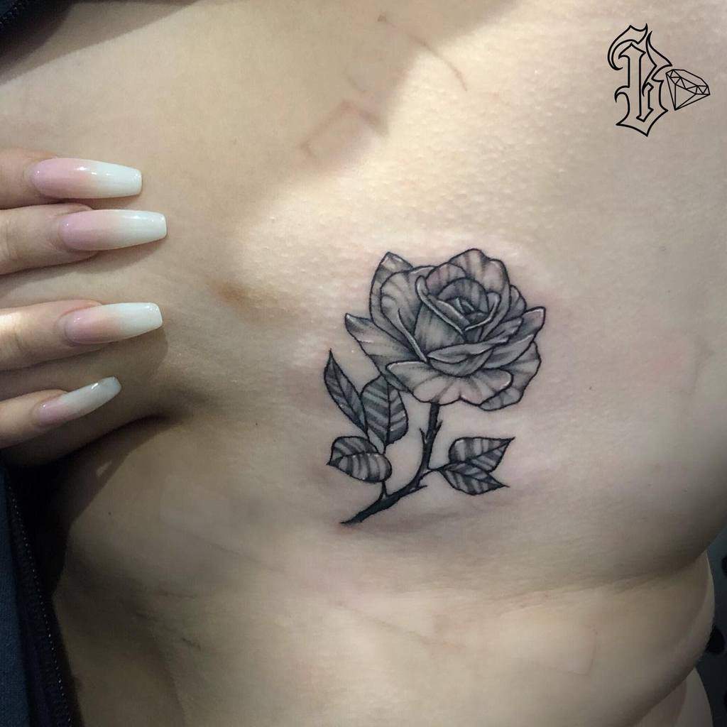 Delicate black and gray rose tattoo with a short stem and leaves, placed subtly on the ribcage area