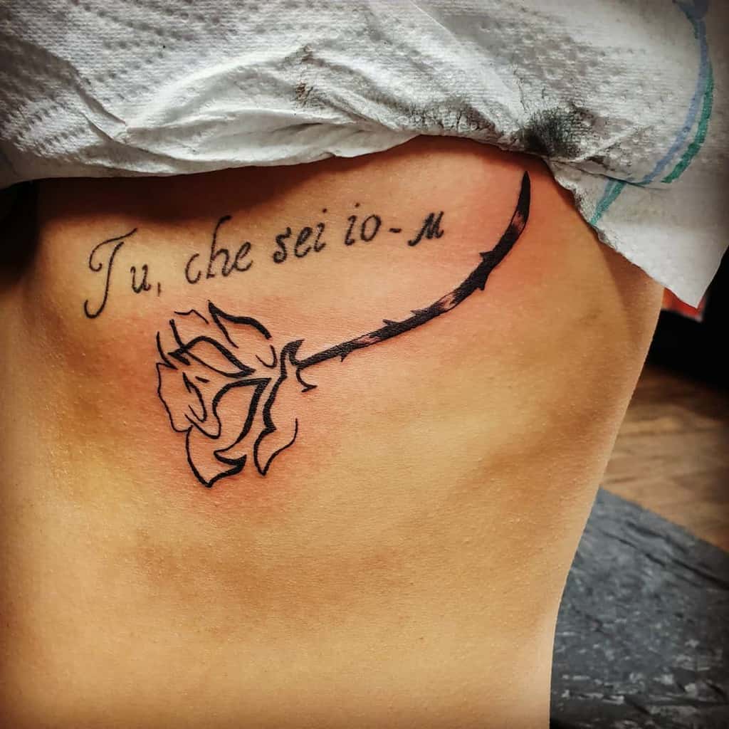 A tattoo featuring a simple black rose with a thorny stem, accompanied by the Italian phrase "Tu, che sei io," on the side of the torso