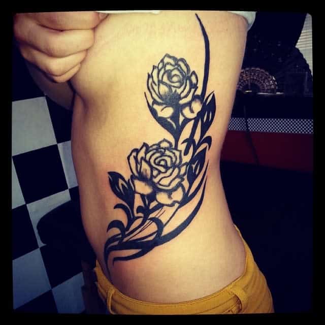 A bold black tribal tattoo of two roses with flowing vines, extending along the side of the torso