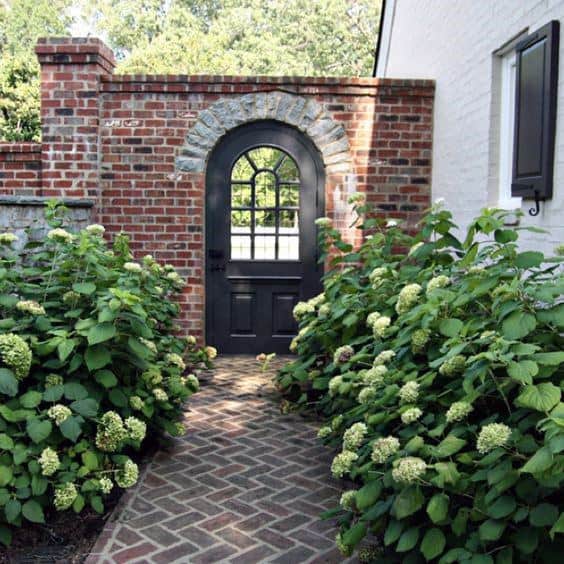 Brick walkway with hydrangeas leading to an arched black door in a brick wall