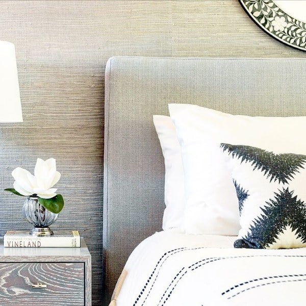 A neatly made bed with patterned pillows, a wooden nightstand, a white lamp, and a vase with a white flower
