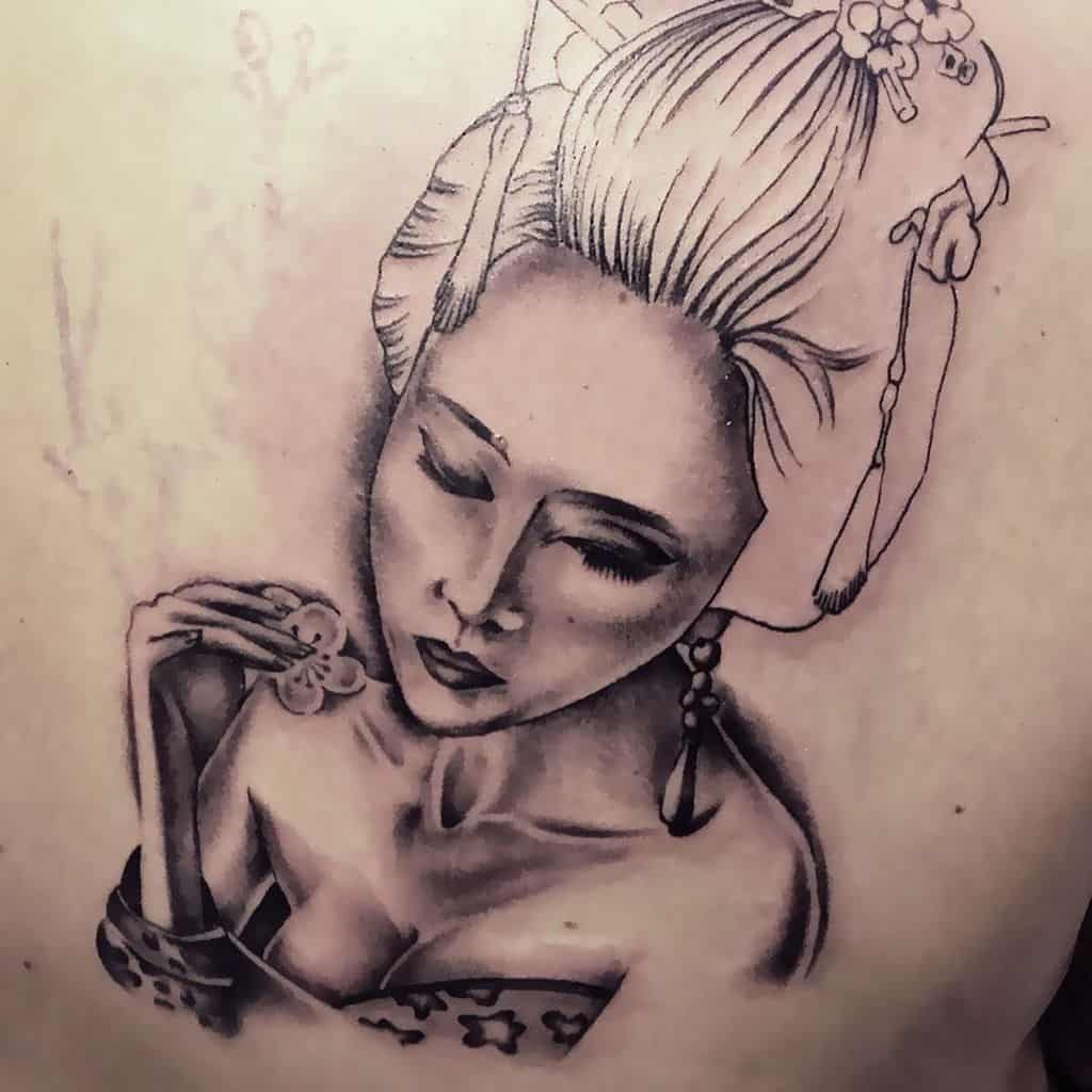 Black and gray tattoo of a serene Geisha with closed eyes, an intricate hairpiece, delicate earrings, and holding a small flower.