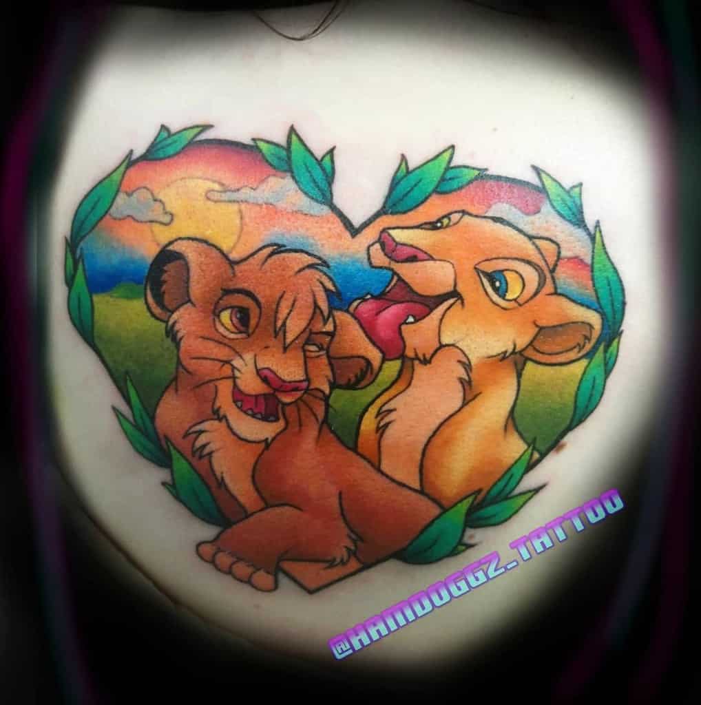 Colorful tattoo of young Simba and Nala, framed within a heart shape and surrounded by greenery and sunset sun with clouds